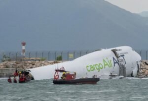 Hong Kong, China: A cargo aircraft operating for Emirates SkyCargo skidded off the north runway at Hong Kong International Airport early Monday, plunging partially into the sea and killing two ground personnel. The four crew members onboard the Boeing 747-400 freighter survived the crash. According to Hong Kong authorities, the accident occurred around 3:50 a.m. local time when the aircraft, identified as TC-ACF, overshot the runway after landing. The plane was operating flight EK9788 from Dubai’s Al Maktoum International Airport on behalf of Emirates SkyCargo. The aircraft is owned and operated by ACT Airlines, a Turkish cargo carrier contracted by Emirates. Initial reports suggest that the aircraft struck a security patrol vehicle positioned near the runway end, forcing the vehicle into the sea. “Both occupants of the vehicle were pulled from the water,” police told Reuters, confirming that one person died at the scene and the other later succumbed to injuries at the hospital. The four Turkish crew members aboard the aircraft were rescued without serious injuries. Photos and videos circulating on social media showed the jet partially submerged near the seawall, with the rear fuselage section visibly damaged. The Airport Authority Hong Kong (AAHK) has temporarily closed the north runway to facilitate investigation and aircraft recovery operations. The airport’s two other runways continue to operate normally. An official investigation has been launched by the Hong Kong Civil Aviation Department. Authorities said they are examining multiple factors, including braking performance, runway conditions, and possible mechanical issues. Early reports indicate that weather conditions were stable at the time of landing, making it unlikely that visibility or wind played a major role. The Boeing 747-400 involved in the accident is 32 years old and had been converted from a passenger aircraft to a freighter several years ago. Emirates confirmed that the flight carried no cargo onboard during the incident. “This tragic incident has claimed the lives of two valued airport workers,” said an Emirates spokesperson, adding that the airline is cooperating fully with local authorities and expressing condolences to the victims’ families.