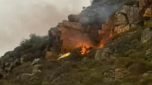 Firefighting Helicopter Crashes In Hout Bay During Wildfire Operations, Pilot Survives SEO DES: The aircraft likely struck terrain during water-dropping operations; the pilot survived with injuries, as crews managed the crash site and secondary fire SOCIAL: A firefighting helicopter crashed over Hout Bay in Cape Town, South Africa on 25 March 2026 while battling a wildfire in Table Mountain National Park. The aircraft likely struck mountainous terrain during water-dropping operations. The pilot survived with injuries, and no fatalities were reported. Emergency teams responded swiftly, managing both the crash site and a secondary fire. The wildfire, which began a day earlier, burned around 160 hectares, prompting trail closures, while authorities have launched an investigation into the incident. Cape Town, South Africa: A firefighting helicopter operating over the slopes of Hout Bay in Table Mountain National Park crashed on the evening of 25 March 2026 while assisting efforts to contain a rapidly spreading wildfire, authorities confirmed. The aircraft, contracted to support firefighting operations coordinated by South African National Parks, went down in mountainous terrain between Hout Bay and Constantia Nek during active aerial suppression efforts. Preliminary reports indicate the helicopter encountered difficulty while conducting water-dropping operations over the fire zone. Officials stated that the aircraft likely clipped the mountainside with its main rotor before crashing. Witness accounts and local authorities reported that the helicopter descended shortly after impact, with flames and smoke visible at the crash site. The pilot survived the crash, with reports consistently confirming that no fatalities occurred. Initial updates indicated the pilot was rescued alive with injuries. Authorities described the survival as remarkable given the challenging terrain and operational conditions. A large-scale emergency response was immediately activated, involving: Wilderness Search and Rescue (WSAR) SANParks Kishugu Aviation Netcare24 City of Cape Town Fire and Rescue Services Local law enforcement units Rescue teams were deployed to access the mountainous crash site, while an Incident Management Team was established to coordinate operations and secure the area. Authorities also managed a secondary fire triggered by the crash, as firefighting operations continued in the surrounding area. The helicopter had been actively engaged in combating a wildfire that ignited on 24 March 2026 along the Hout Bay slopes. The blaze spread across mountainous terrain between Hout Bay and Constantia Nek Approximately 160 hectares of vegetation were affected Strong winds contributed to flare-ups and fire spread Multiple aerial assets, including helicopters and fixed-wing aircraft, had been deployed to support ground firefighting crews. As a precaution, authorities closed hiking trails and access routes in the affected areas to ensure public safety and allow uninterrupted firefighting operations. SANParks confirmed that an investigation has been launched to determine the exact cause of the crash. In an official statement, the agency said it is unable to provide further details at this stage as inquiries remain ongoing.