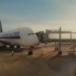Singapore: Singapore Airlines (SIA) Group reported a sharp 57.4 per cent decline in annual net profit for the financial year ended March 31, 2026, as mounting losses at Air India and the absence of a one-time accounting gain from the Vistara merger weighed heavily on the carrier’s bottom line. The airline group posted a net profit of SGD 1.184 billion for FY2025/26, down from SGD 2.778 billion recorded a year earlier. Despite the steep fall in earnings, SIA’s core airline operations remained strong, with the group posting record revenue and higher operating profit driven by sustained passenger demand and network expansion. According to SIA’s earnings report, the biggest factor behind the profit decline was the absence of the SGD 1.098 billion non-cash accounting gain booked in November 2024 following the completion of the Air India-Vistara merger. The previous year’s results had been significantly boosted by that one-off gain after SIA’s stake in Vistara was converted into a 25.1 per cent shareholding in the enlarged Air India Group. At the same time, SIA swung from reporting profits from associated companies last year to posting a SGD 846 million share of losses in FY26, mainly due to Air India’s worsening financial performance. The airline said this year’s accounts reflected a full year of Air India losses, compared to only four months in the previous financial year after the merger completion. Financial disclosures from Singapore Airlines indicated that Air India Group, comprising Air India and Air India Express, recorded losses of approximately SGD 3.56 billion, equivalent to around USD 2.8 billion or more than ₹26,000 crore during FY2025/26. Some reports estimated the losses could be even higher at nearly ₹28,300 crore depending on accounting adjustments and currency conversion methods. The losses mark Air India’s largest annual financial setback since the Tata Group reacquired the airline in 2022. Air India has not yet officially released its standalone FY26 financial statements. Singapore Airlines attributed Air India’s financial pressure to multiple operational and geopolitical challenges, including elevated jet fuel prices, Middle East airspace restrictions, supply-chain disruptions, aircraft delivery delays, and the continued closure of Pakistani airspace for Indian airlines. These factors forced Air India to reroute several long-haul flights, increasing fuel burn and operating costs. The impact of the Iran conflict and the closure of the Strait of Hormuz also emerged as a major concern for SIA itself. The airline warned that higher jet fuel prices resulting from the conflict are expected to weigh more heavily on earnings in the coming fiscal year because fuel costs are reflected on a lagged basis. Air India recently announced significant reductions to its international operations between June and August 2026, including suspending services on several overseas routes and reducing frequencies to destinations in Europe and North America. Routes affected include Delhi-Chicago, Mumbai-New York, and Delhi-Shanghai, while services to Paris, Milan, and Rome are also being reduced. Despite the financial turbulence, Singapore Airlines reiterated its long-term commitment to Air India and described the investment as a “core component” of its multi-hub strategy. The group said its partnership with Tata Sons provides direct access to one of the world’s fastest-growing aviation markets while complementing SIA’s Singapore hub operations. SIA stated that Air India continues to make progress in its fleet renewal and aircraft retrofit programme, customer experience initiatives, and operational performance improvements despite the difficult environment. While net profit fell sharply, Singapore Airlines’ operational performance remained resilient. Group revenue rose five per cent year-on-year to a record SGD 20.55 billion, supported by strong passenger demand, higher yields, and robust travel flows across Asia and Europe. Operating profit climbed 39 per cent to approximately SGD 2.52 billion, and the airline carried a record 42.4 million passengers during the year. The airline also maintained expansion plans despite industry pressures. Reuters reported that SIA intends to continue adding capacity and launching new services, including flights connecting Singapore and Madrid via Barcelona, while increasing frequencies to Manchester, Milan, Munich, and London Gatwick.